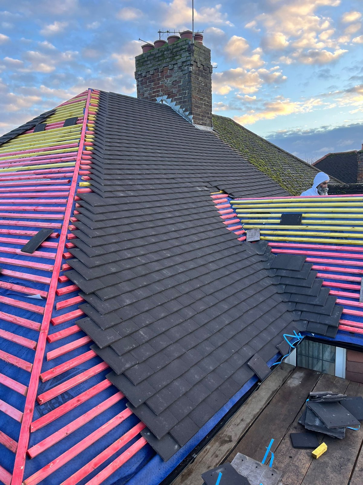 Roofer in safety gear working on a slate roof with colorful neon pink and lime striped paint patterns, chimney and sky visible